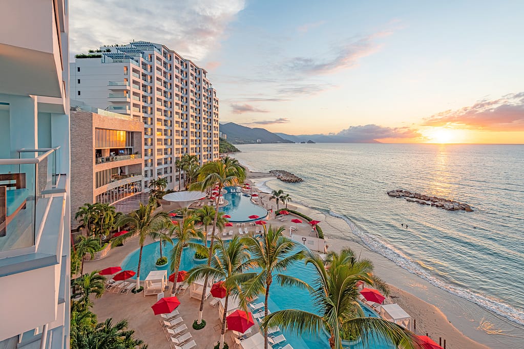 A sunset view of a beach resort with a large pool area, palm trees, and lounge chairs with red umbrellas facing the ocean.