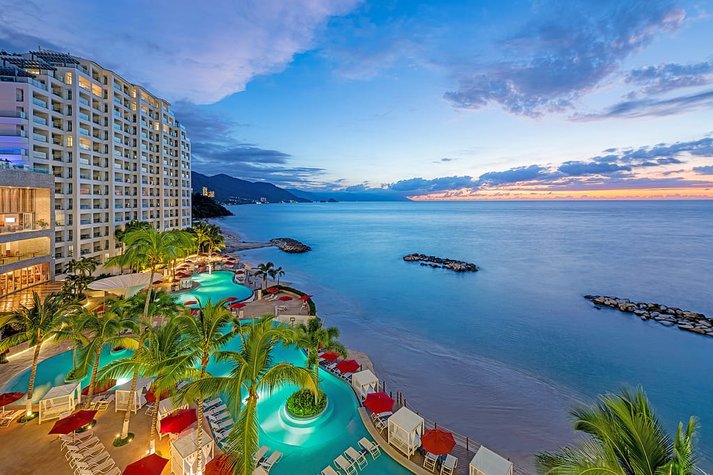 A stunning view of a Hilton resort at sunset, showcasing modern architecture, vibrant pools, and the ocean in the background.