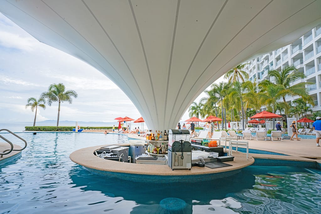 View of a luxurious pool area featuring a swim-up bar, surrounded by sun loungers and palm trees, with distant guests enjoying the resort atmosphere.