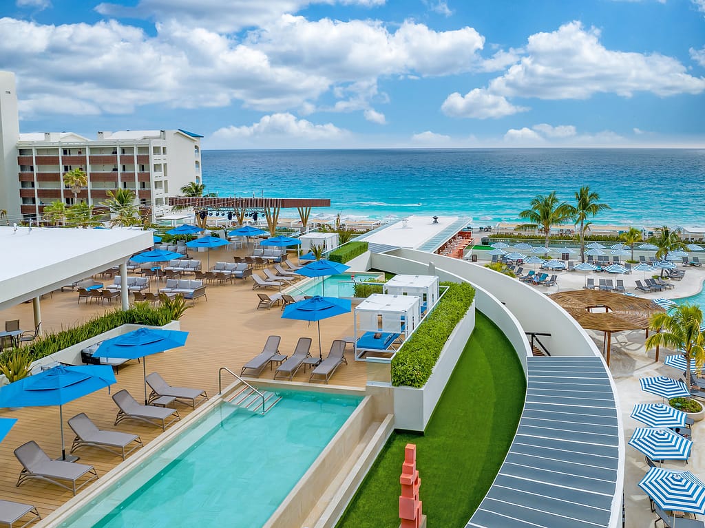 Aerial view of a modern resort with a swimming pool, sun loungers, and blue umbrellas overlooking a beautiful ocean landscape.